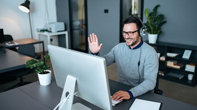 Ein junger Mann sitzt im Büro und hebt die Hand lachend zum Gruß vor der Webcam