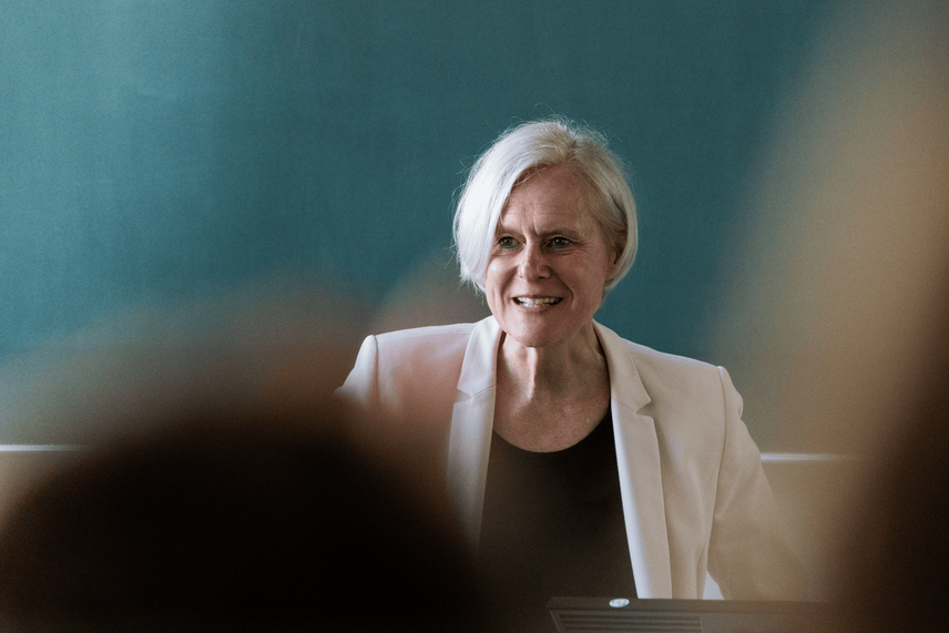 Eine sprechende Frau in einem hellen Blazer, die vor einer grünen Tafel steht