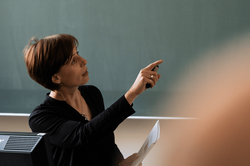 Eine Frau die auf etwas zeigt und vor einer grünen Tafel steht