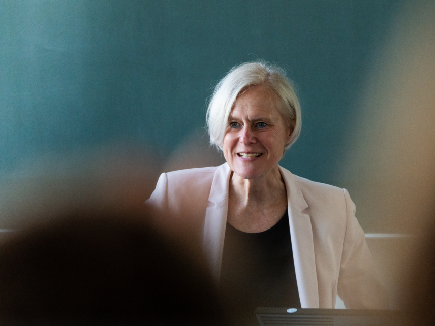 Eine sprechende Frau in einem hellen Blazer, die vor einer grünen Tafel steht