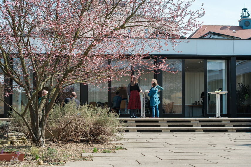 Menschen, die auf einer Terasse stehen. Im Vordergrund eine blühende Zierkirsche.