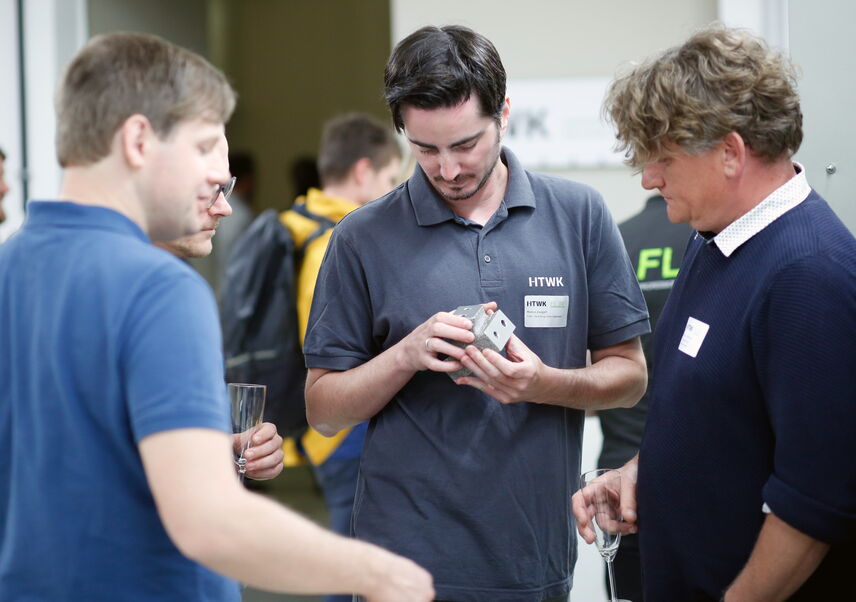 Drei Personen betrachten ein kleines Bauteil aus Metall, dass die mittlere Person in den Händen hält