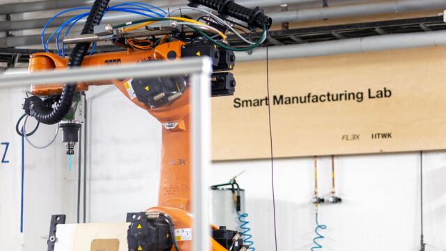Teilausschnitt des Smart Manufacturing Lab im Vordergrund ist ein Teil einer Maschine in Orange zu sehen und im Hintergrund das Namensschild des Labs