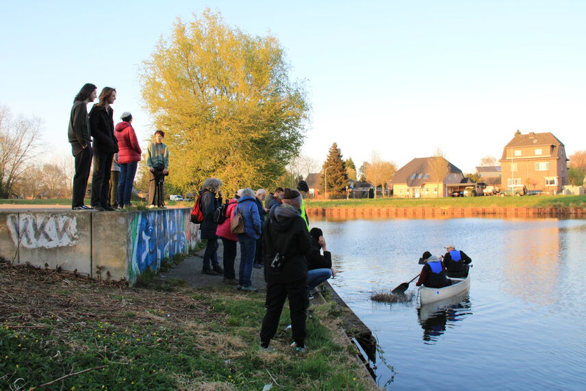 2 Personen im Paddelboot mit angehängter bepflanzter Schwimminsel am Ufer, an welchem sich Zuschauende befinden