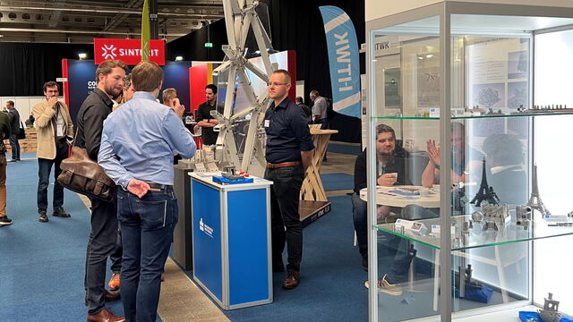 Es ist der Stand der Hochschulen zu sehen. An einem Pult stehen drei Männer, die in einem Gespräch sind. Im Hintergrund sieht man den Demonstrator, im Vordergrund steht eine Vitrine mit Exponaten.