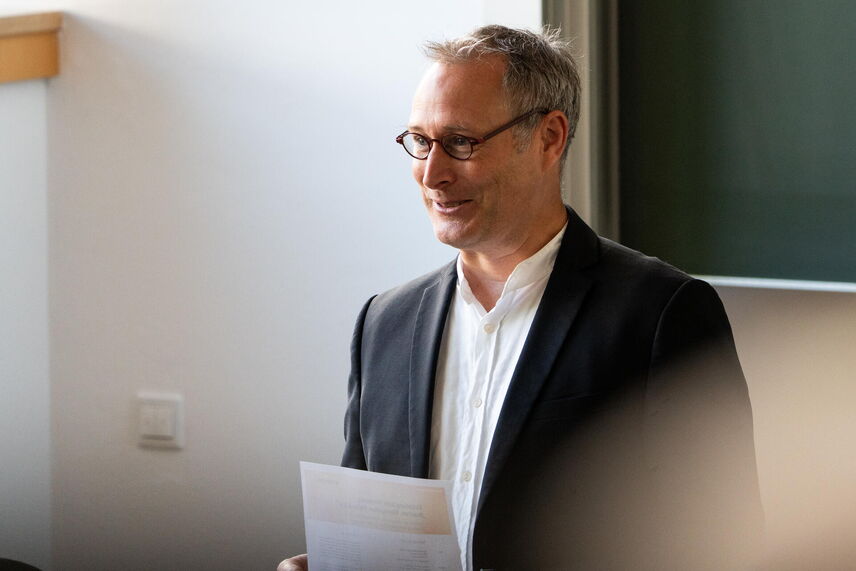 Ein sprechender Mann mit Brille, in einem schwarzen Jacket, vor einer grünen Tafel