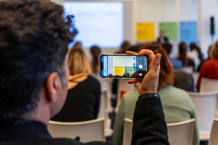 Ein Mann hält ein Handy in der Hand. Im Hintergrund ist unscharf das Publikum zu erkennen.