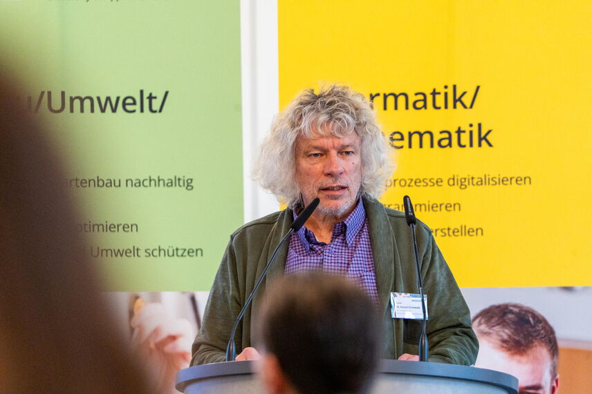 Ein sprechender Mann, der vor einer Wand mit bunten Postern steht und von Menschen umgeben ist.