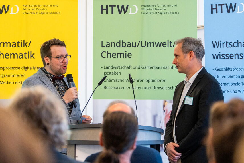 Zwei Männer stehen vor einer bunten Fläche. Im Vordergrund unscharfe Hinterköpfe des Publikums