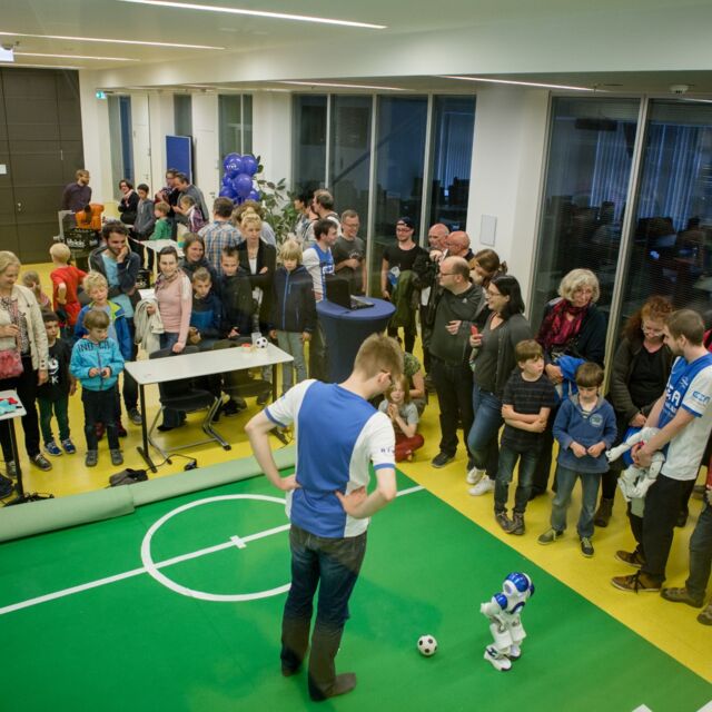 Viele Personen stehen um ein kleines Fußballfeld auf dem ein Roboter gesteuert wird.