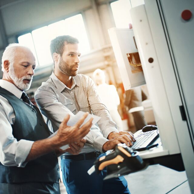 Eine ältere männliche Person mit grauen Haaren und eine jüngere Person stehen zusammen an einem Arbeitsplatz und betrachten ein Blatt und einen Bildschirm.