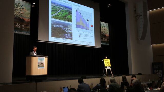 Ein Mann der an einem Rednerpult in einem dunklen Raum steht. Daneben eine hellprojizierte Fläche mit Bildern und Diagrammen.