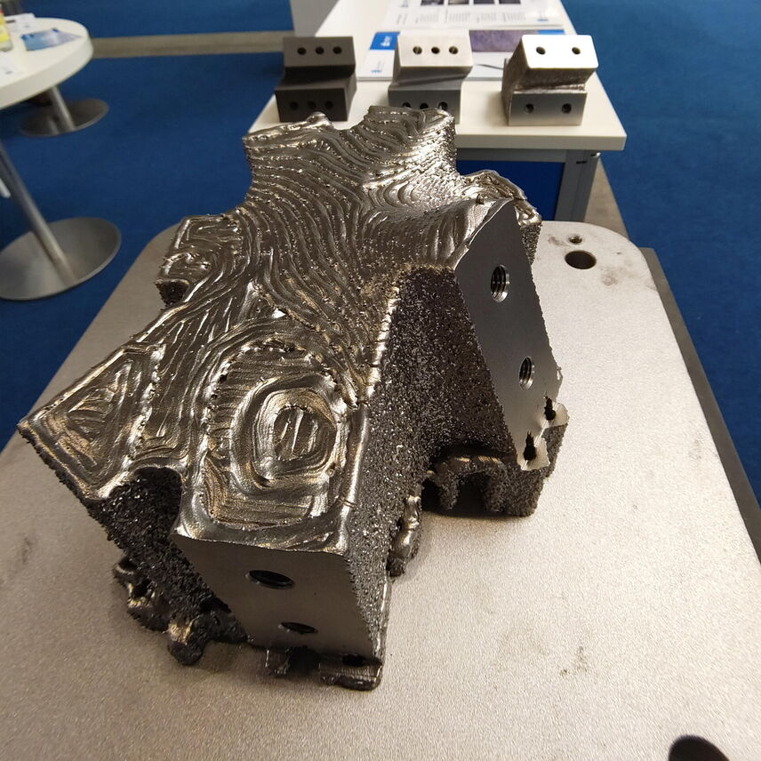 Auf dem Tisch liegt ein Bauteil aus Metall, welcher auch Knoten genannt wird. Er hat eine unebene Oberfläche und Löcher an den Seiten.