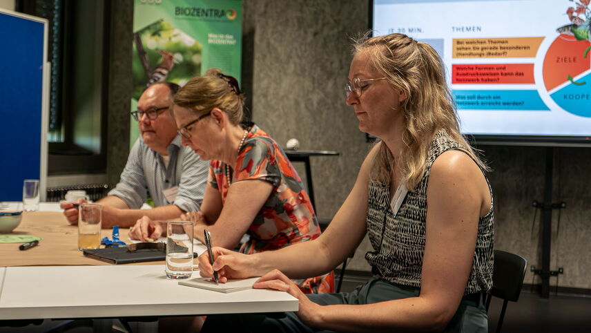 Eine Frau, die mit anderen Teilnehmenden an einem Tisch sitzt und etwas aufschreibt.