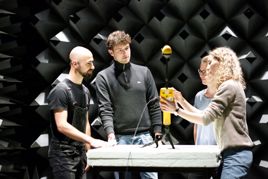 Vier Menschen stehen in einem Raum mit schwarzen pyramidenförmigen Strukturen an der Wand und betrachten einen gelben Apparat der mittig auf einem Tisch platziert ist