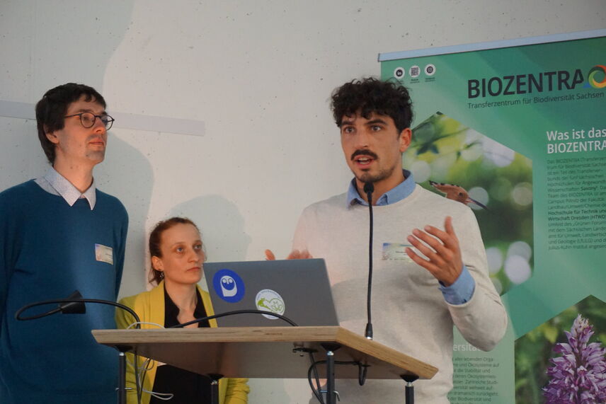 Ein Mann, der neben zwei Personen steht und einen Vortrag hält.