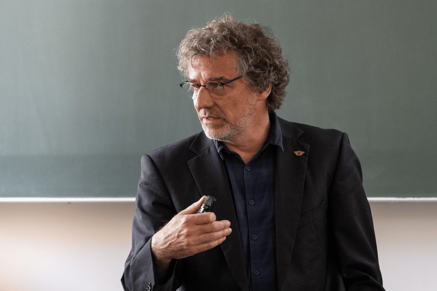 Ein sprechende Mann in einem schwarzen Jacket, vor einer grünen Tafel