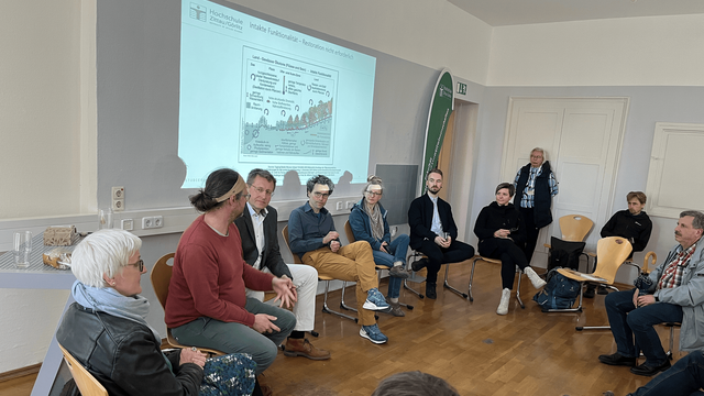 Teilnehmende einer Podiumsdiskussion sitzen im Stuhlkreis in einem Seminarraum der ehemaligen Baugewerkeschule in Zittau. Im Hintergrund ist eine Präsentation mit dem Logo der Hochschule Zittau/Görlitz zu sehen.