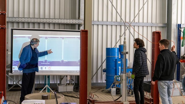 Eine Person erklärt Messdaten auf einem Bildschirm, während zwei Personen rechts im Bild zuhören und eine Versuchsanlage im Geotechnikum beobachten