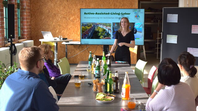 Maria Fabisch spricht vor einem Bildschirm zu mehreren an einem Tisch sitzenden Zuschauer*innen.