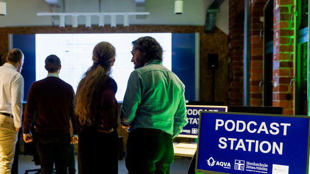 Mehrere Personen stehen vor einer großen Leinwand, auf Bildschirmen steht "Podcaststation"