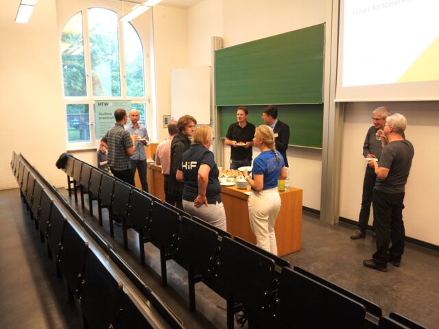 Mehrere Personen stehen um einen Tisch in einem Hörsaal, essen und unterhalten sich.