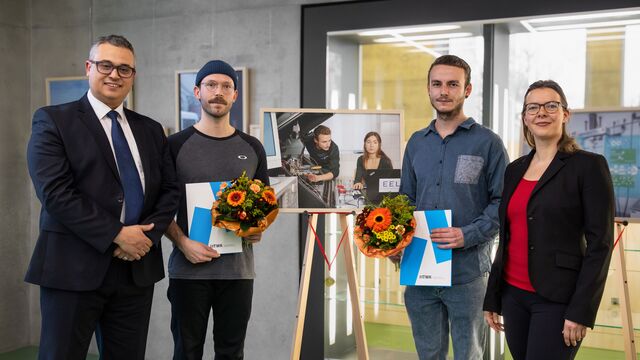 Drei Männer und eine Frau stehen vor einer Staffelei mit gerahmtem Bild, zwei Männer halten Urkunden und Blumen in den Händen