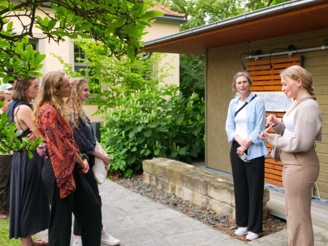 Zwei Frauen, die vor einem Gartenhaus stehen und zu einer gruppe Menschen sprechen