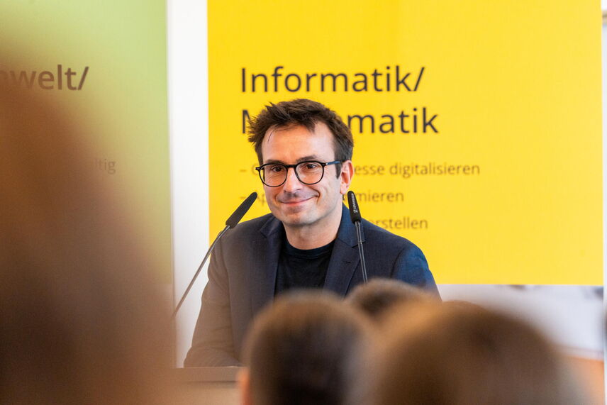 Ein sprechender Mann, der vor einer Wand mit bunten Postern steht und von Menschen umgeben ist.