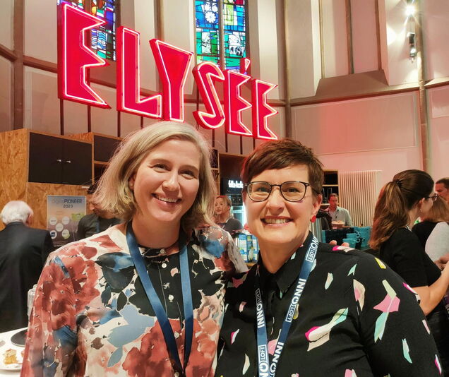 zwei Frauen lächeln in die Kamera, um sie herum stehen viele Menschen