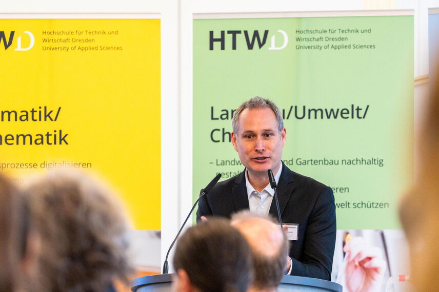 Ein sprechender Mann, der vor einer Wand mit bunten Postern steht und von Menschen umgeben ist.