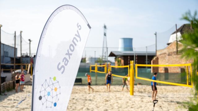 Personen stehen auf einem Beachvolleyball-Feld und im Vordergrund steht eine Beach Flag.