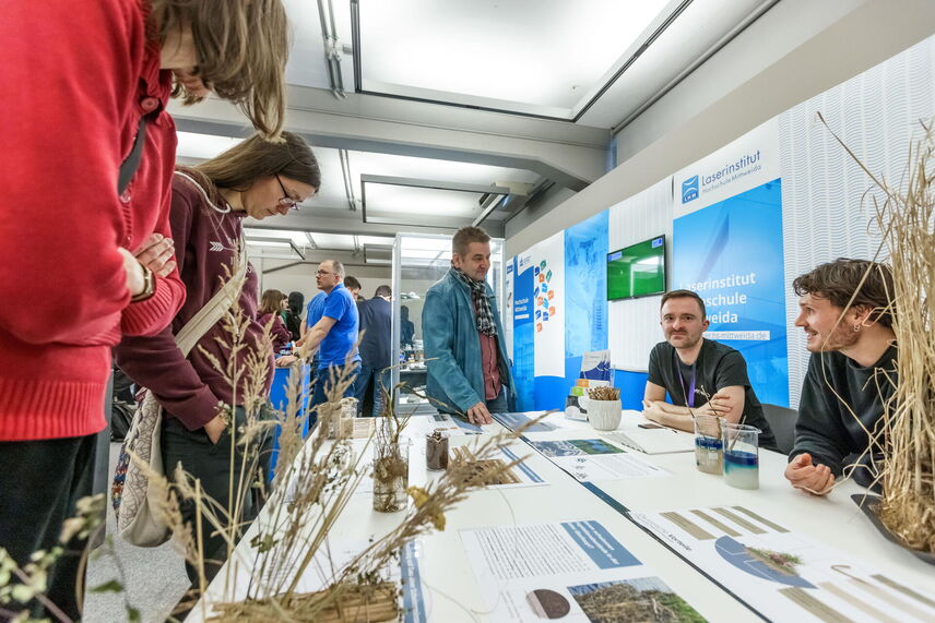 Zwei Forschende der HTWD sitzen am Stand und erklären Besuchenden die Exponate, die auf dem Tisch zu sehen sind.