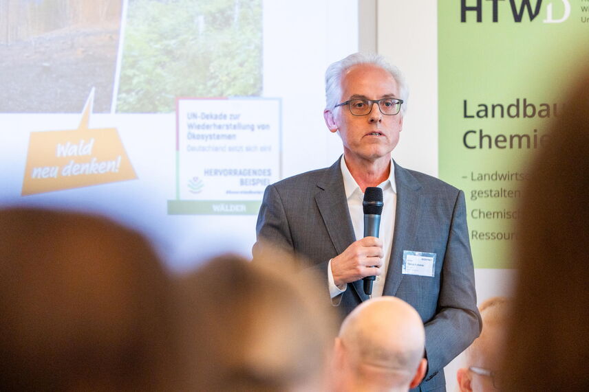 Ein sprechender Mann, der vor einer Wand mit bunten Postern steht und von Menschen umgeben ist.