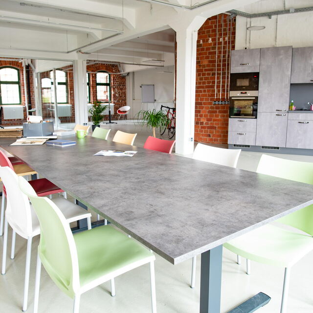 Blick in ein großes Arbeitsloft mit Backsteinwänden mit langem Tisch und vielen Stühlen in verschiedenen Farben.