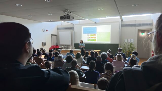 Viele sitzende Personen, die zu einer Frau sehe, die hinter einem Pult, vor einer bunt beleuchteten Fläche steht.