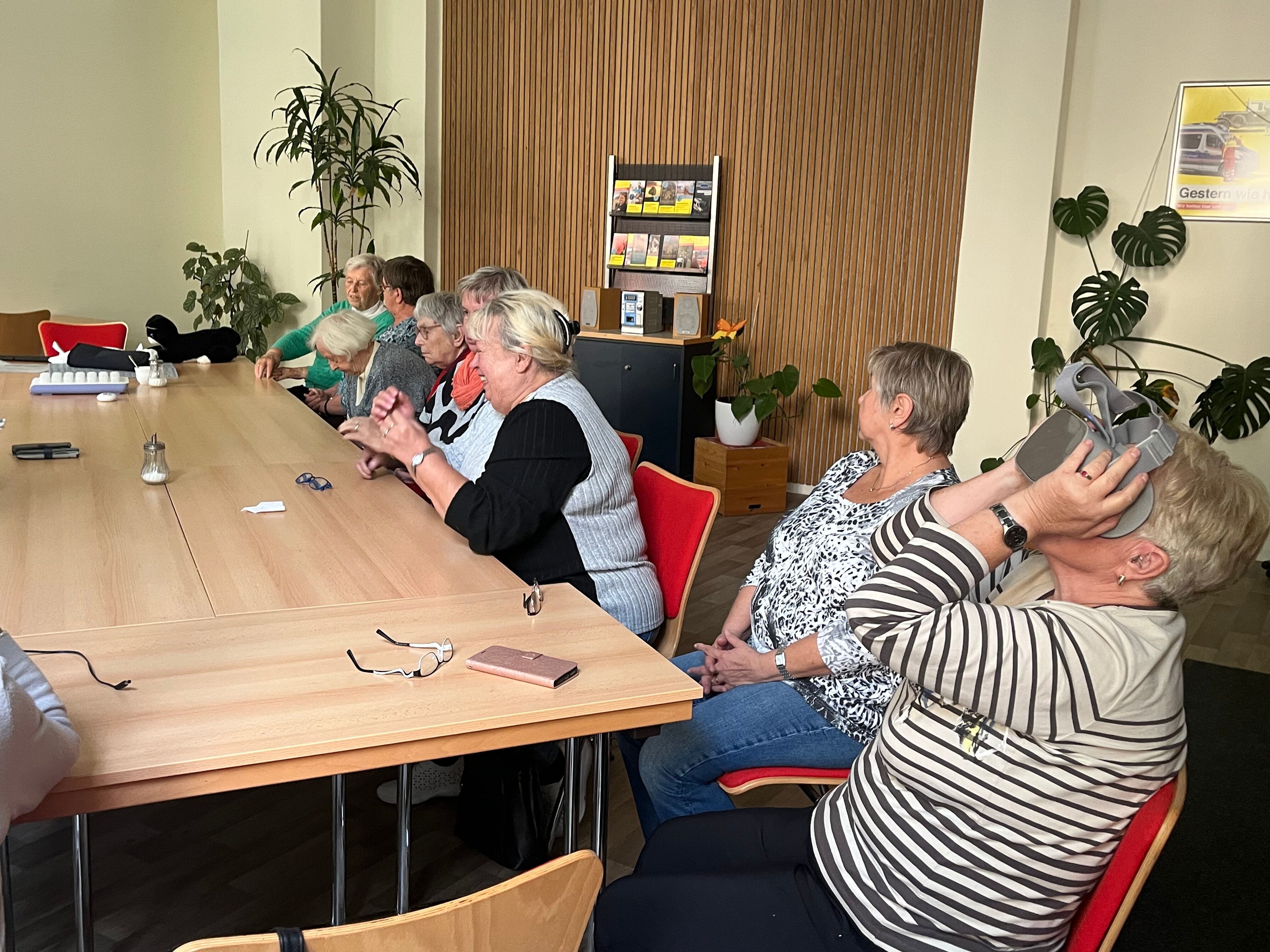 Seniorinnen sitzen zusammen an einem Tisch, eine von ihnen probiert eine VR-Brille aus