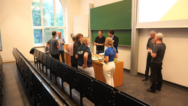 Mehrere Personen stehen um einen Tisch in einem Hörsaal, essen und unterhalten sich.