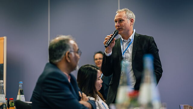 Ein Mann der in ein Mikrofon spricht. Im Hintergrund sitzen Menschen an Tischen.