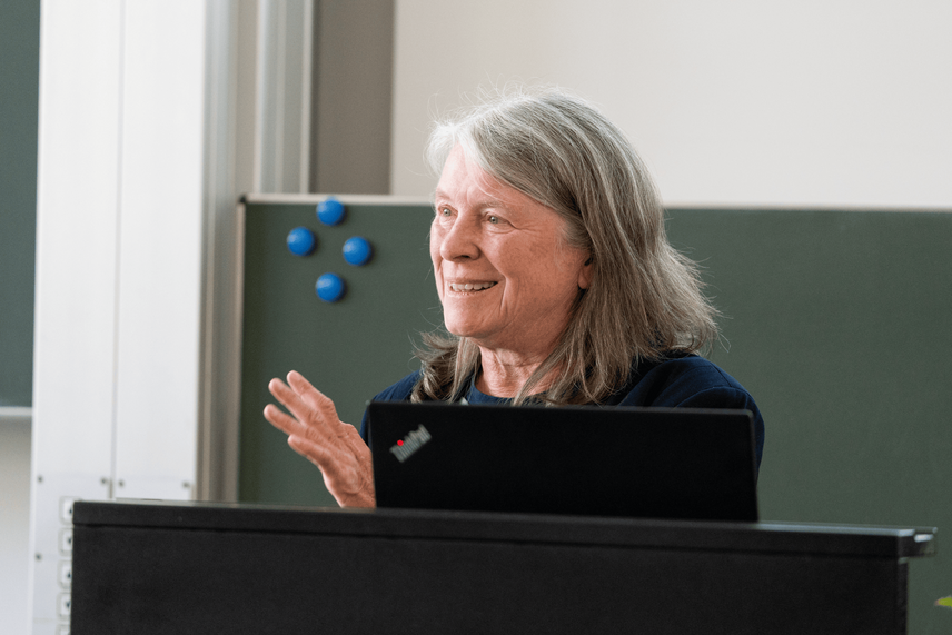 Ein Frau die spricht, vor einer grünen Fläche, mit schwarzen Flächen im Vordergrund