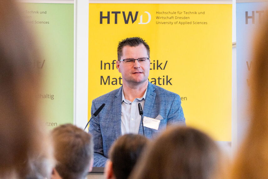 Ein sprechender Mann, der vor einer Wand mit bunten Postern steht und von Menschen umgeben ist.