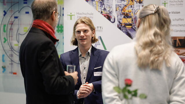 Zwei Männer unterhalten sich, eine Frau hält eine Rose hinter dem Rücken.