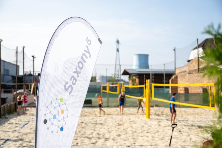 Personen stehen auf einem Beachvolleyball-Feld und im Vordergrund steht eine Beach Flag.