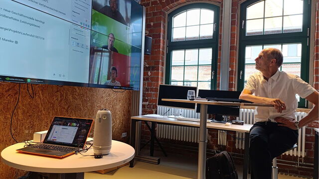 Eine Person sitzt vor einem großen Bildschirm, auf dem mehrere Personen in einem Videocall sind.