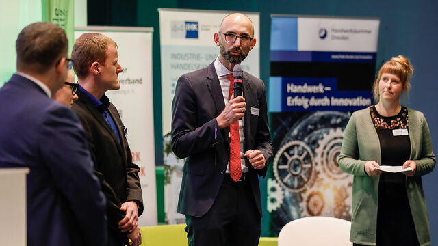 Fünf Personen stehen auf der Bühne, ein Mann spricht ins Mikrophon