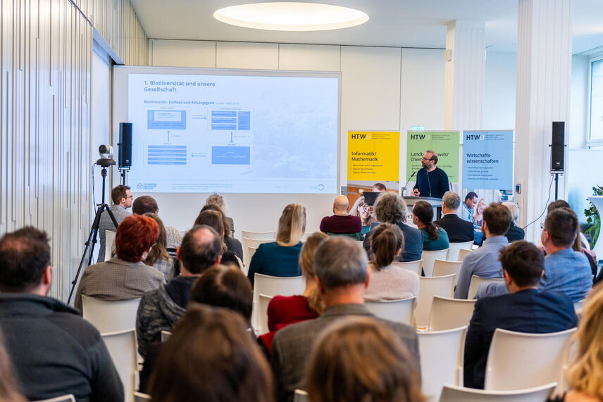 Ein sprechender Mann, der vor einer Wand mit bunten Postern steht. Im Vordergrund sitzen viele Menschen.