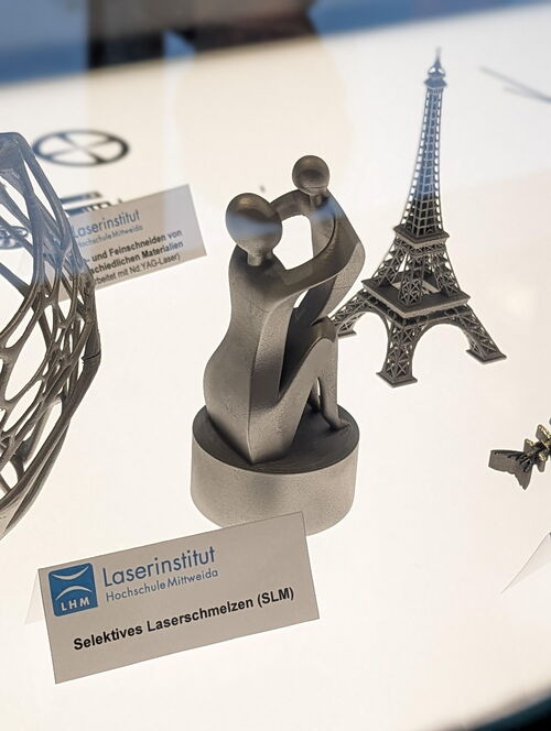 Im Vordergrund steht eine kleine Figur, die eine Frau mit einem Kind zeigt. Im Hintergrund steht der Eifelturm in Miniaturformat. Beide Figuren sind aus Metall und durch den 3D-Druck entstanden.