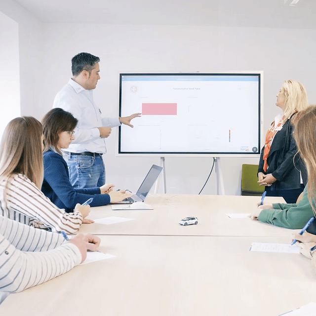 Zwei Personen halten eine Präsentation an einem Bildschirm, im Vordergrund sitzen mehrere Personen an einem Tisch und hören zu.