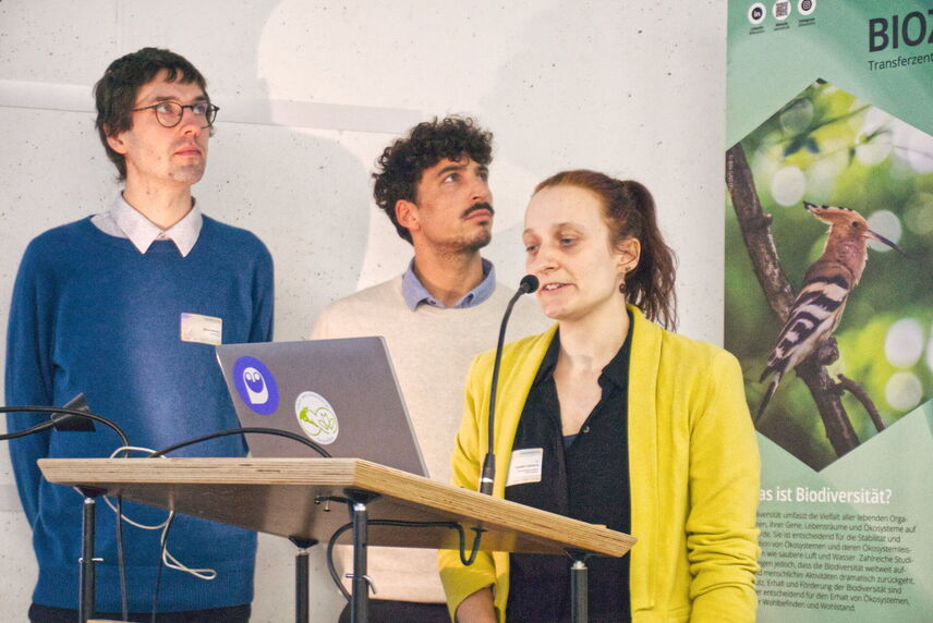 Eine Frau, die neben zwei Männern steht und einen Vortrag hält