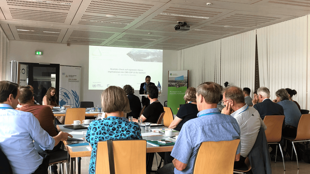 Menschen an Gruppentischen die einem Mann vor einer hell projezierten Leinwand zuhören